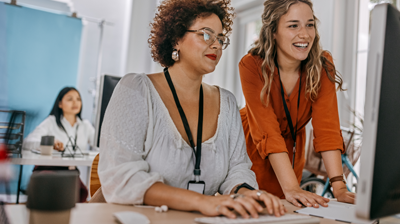 EZLynx Street Smart Webinar Three women in a office at their desks. One woman is sitting at her desk using her computer while another woman in leaning on the desk and looking at the same computer